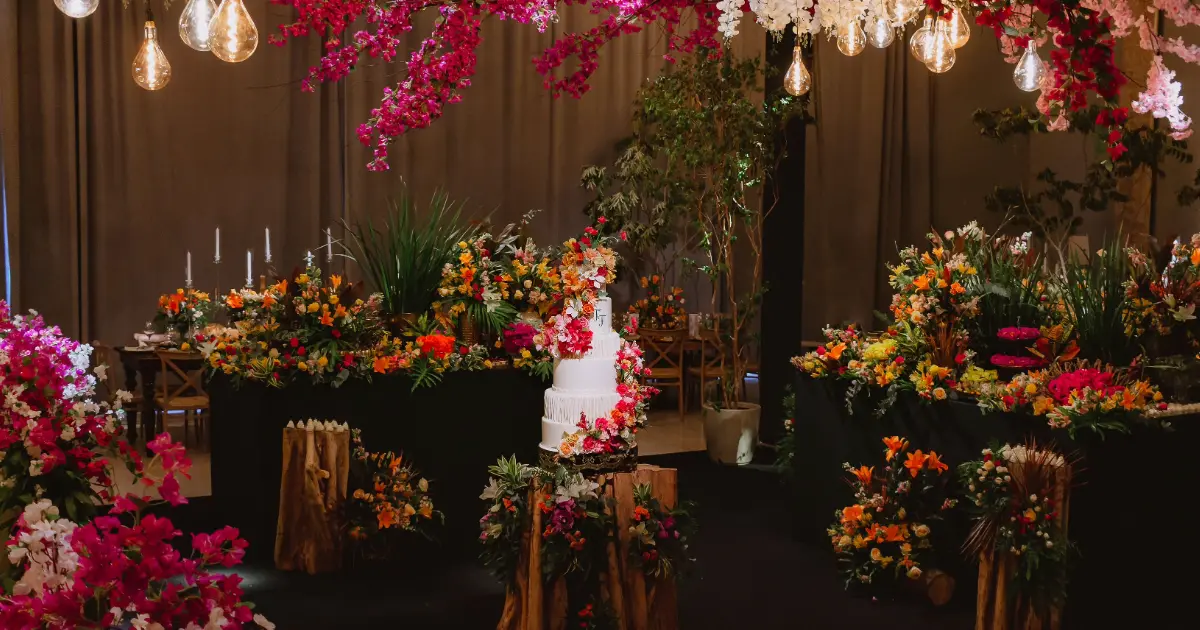 Dekorasi pernikahan indoor dipenuhi rangkaian bunga berwarna-warni, kue pengantin, lampu gantung, dan meja berornamen kayu.