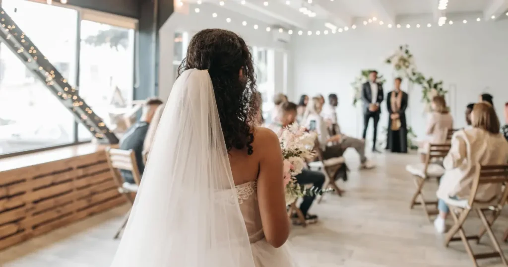 Pengantin berjalan menuju altar dalam ruangan dengan kursi kayu, bunga, dan lampu sebagai dekorasi pernikahan sederhana.