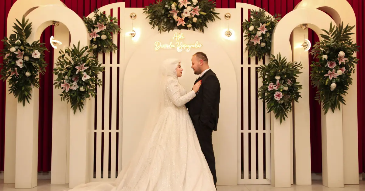 Pasangan pengantin berdiri di depan pelaminan indoor dengan rangkaian bunga dan dekorasi lengkung berwarna putih.