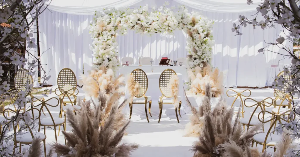 Dekorasi pernikahan outdoor dengan rangkaian bunga putih, kursi emas, pampas, dan altar di area terbuka.