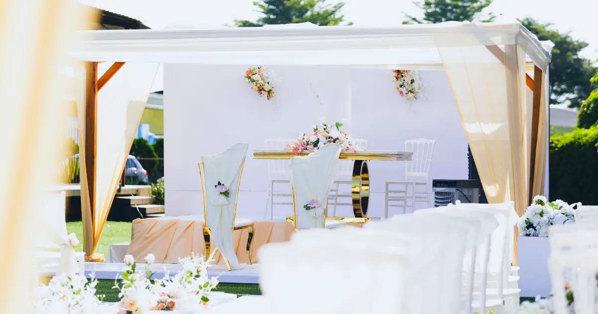Dekorasi pernikahan outdoor dengan kursi putih, bunga, meja pengantin, dan kanopi di area terbuka.