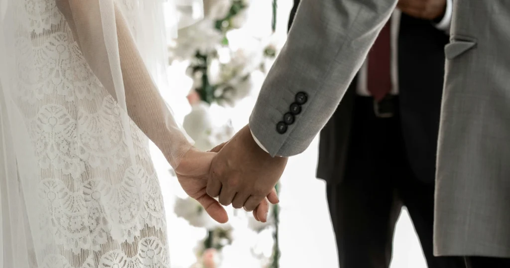 Pasangan pengantin bergandengan tangan dalam suasana pernikahan dengan dekorasi bunga putih di belakangnya.