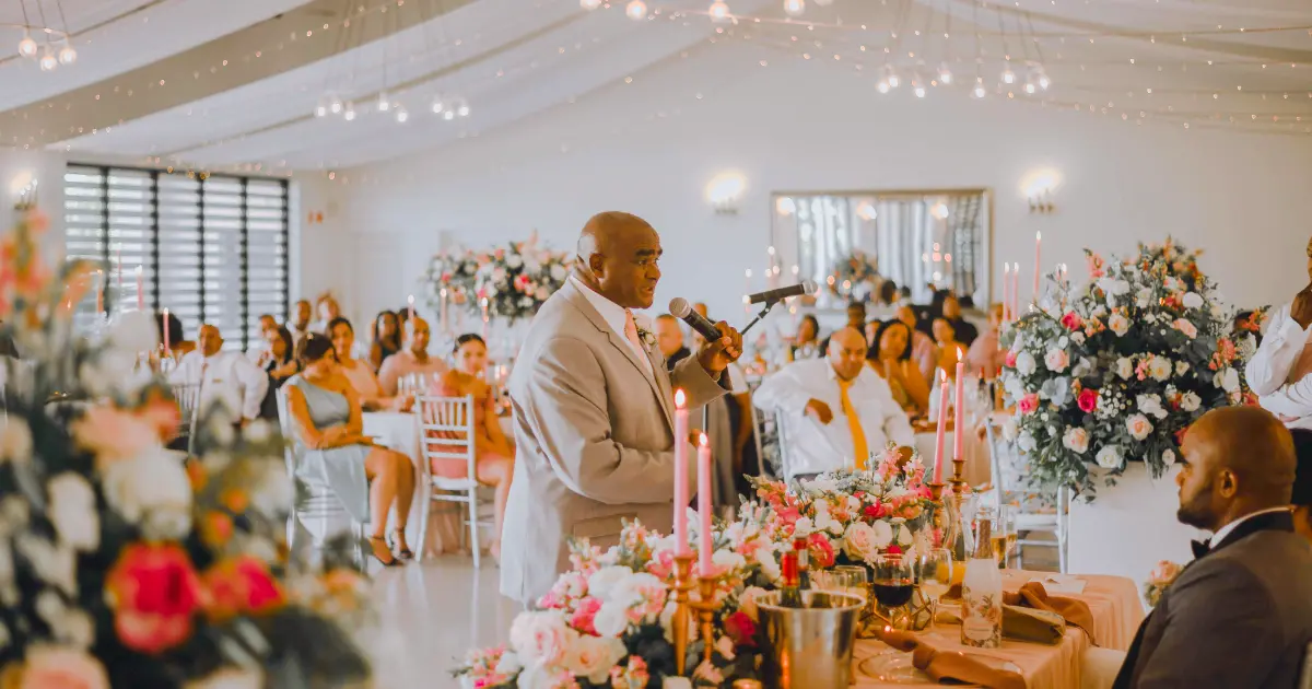 Pengantin dan tamu menghadiri pernikahan indoor dengan pembicara memegang mikrofon di tengah dekorasi bunga.
