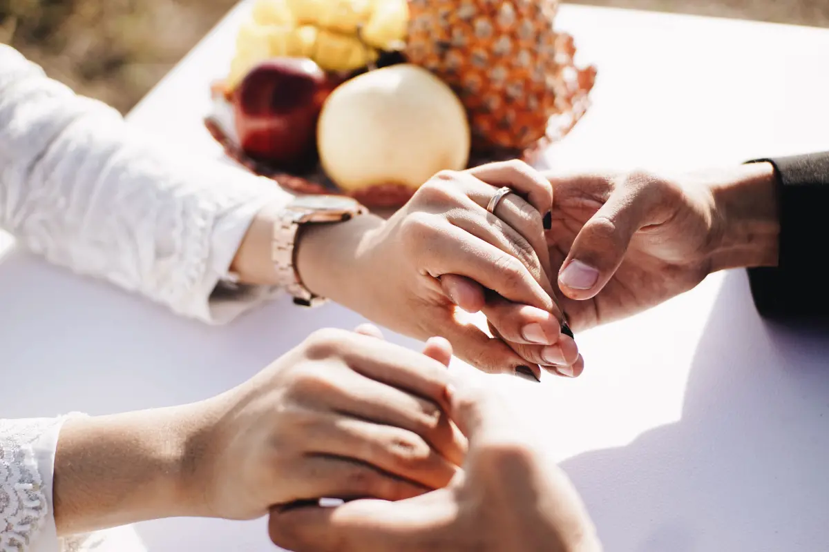 Pasangan mengenakan cincin pernikahan dengan tangan saling menggenggam di meja dalam suasana acara pernikahan.