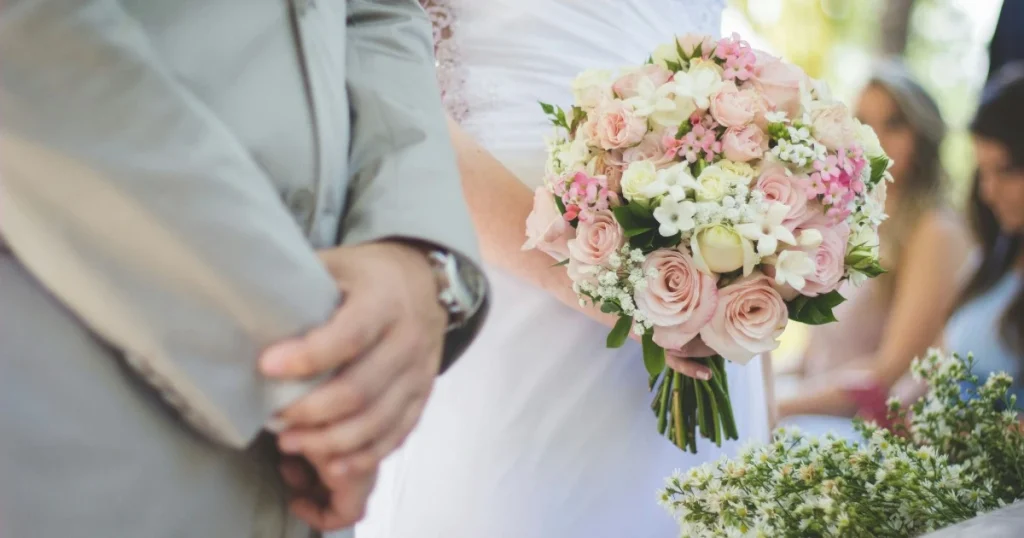 Pengantin membawa buket bunga merah muda dan putih sambil berdiri di samping pasangan dalam acara pernikahan.