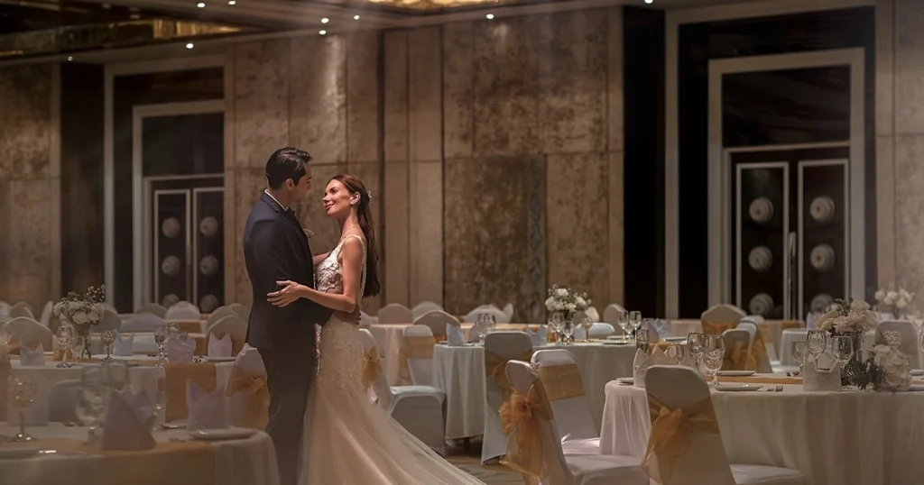 Pengantin berdiri di tengah ballroom dengan meja makan tertata untuk resepsi pernikahan yang elegan.