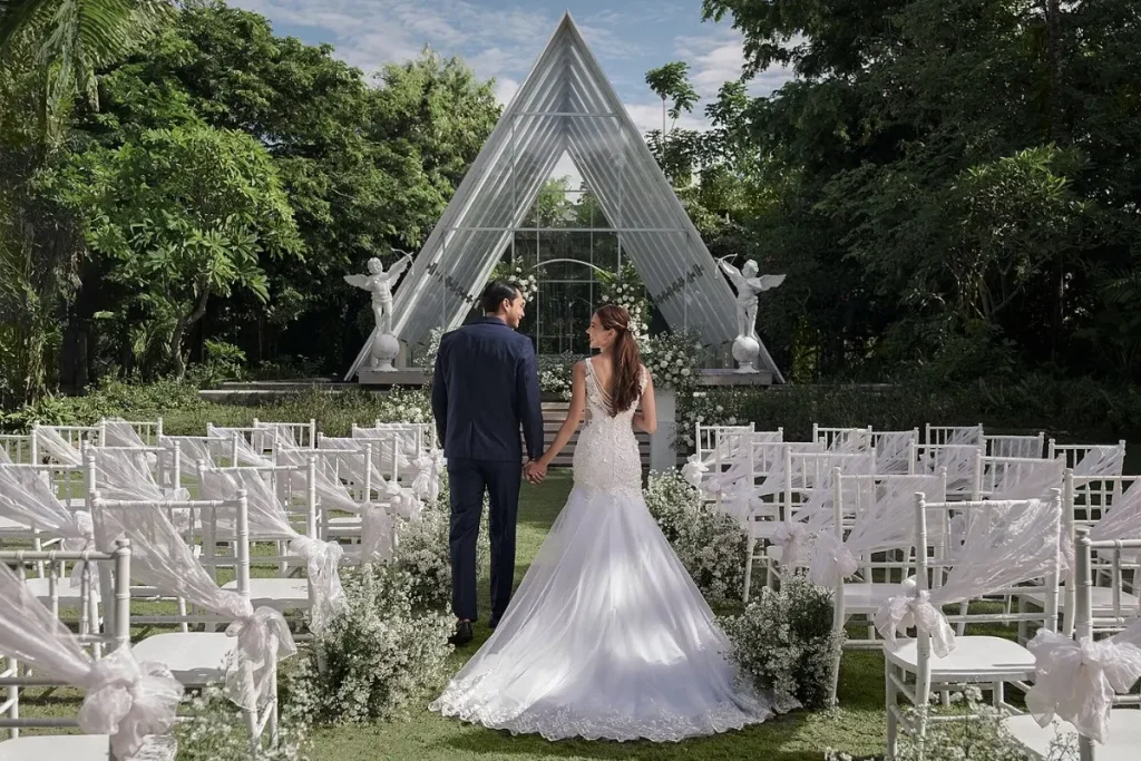 Pasangan pengantin berjalan menuju altar di taman dengan kursi tamu dan dekorasi bunga putih.