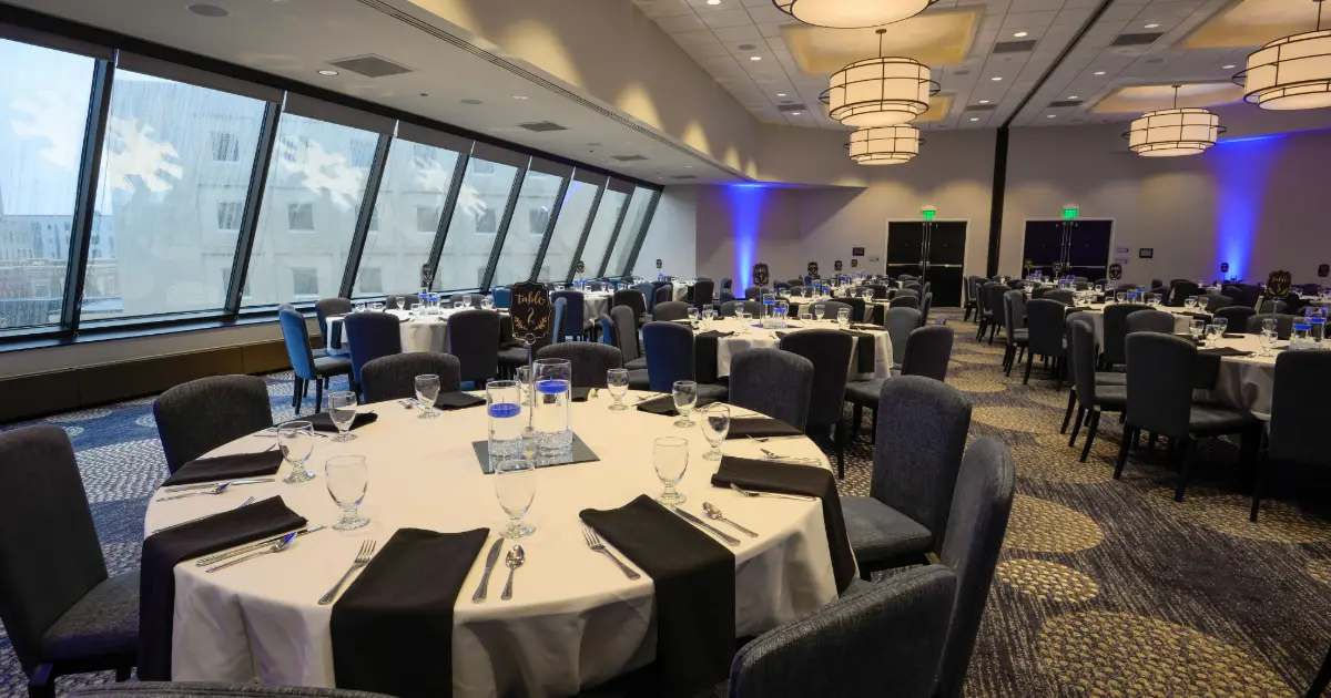 Ballroom menampilkan banyak meja bundar dengan kursi dan perlengkapan makan untuk acara rapat atau pertemuan.
