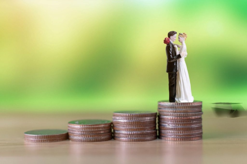 Miniatur pasangan pengantin berdiri di atas tumpukan koin dengan ketinggian berbeda, menggambarkan perencanaan biaya intimate wedding di Bali.