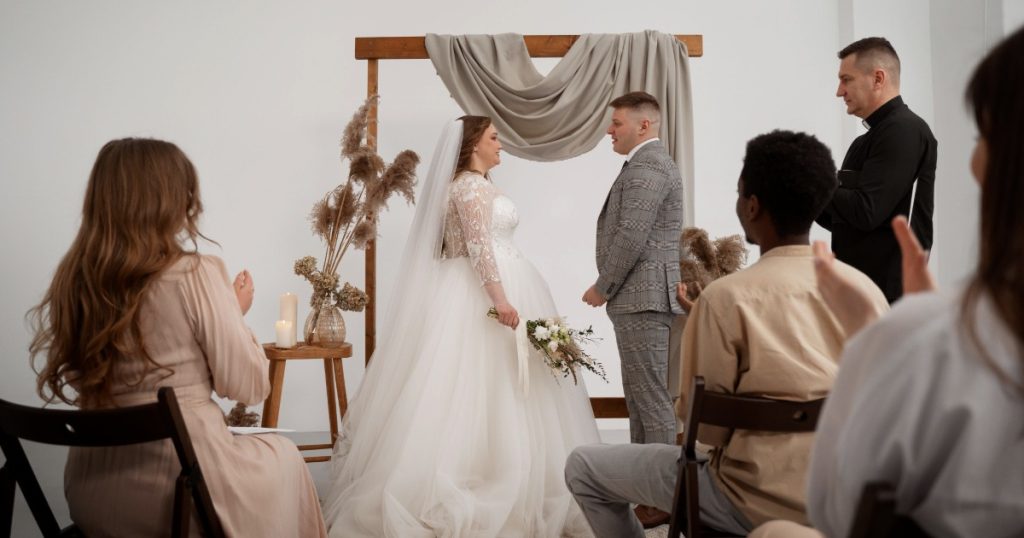 Pasangan pengantin berdiri di depan altar sederhana dengan dekorasi kain, bunga kering, dan beberapa tamu dalam intimate wedding.