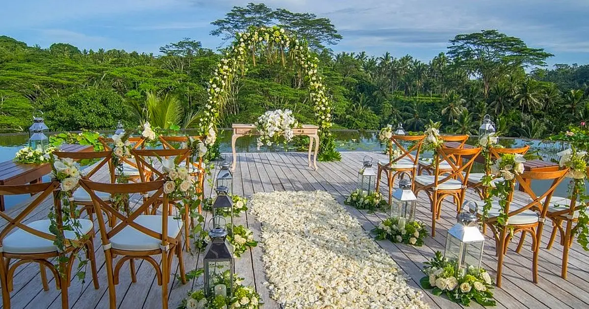 Area pernikahan di atas kolam dengan kursi kayu, lorong bunga putih, dan pemandangan hijau di sekitarnya.