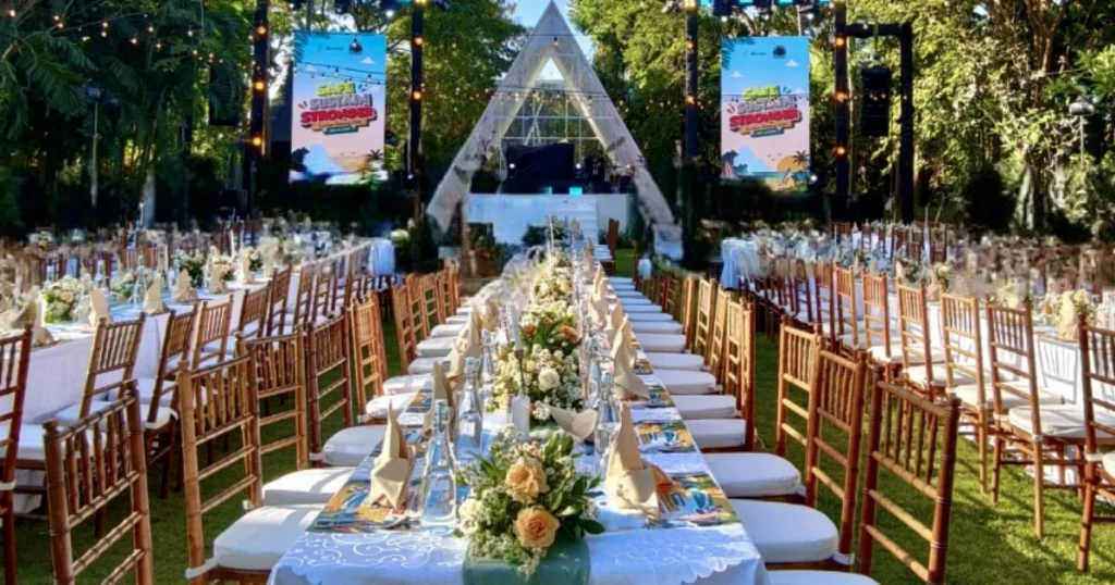 Meja panjang dengan dekorasi bunga, kursi tertata, dan panggung di taman untuk acara gala dinner perusahaan di Bali.