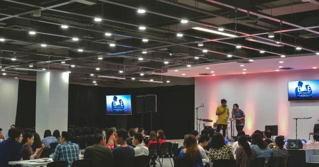 Peserta menghadiri acara di dalam venue dengan pembicara di panggung, layar presentasi, mikrofon, dan peralatan audio.