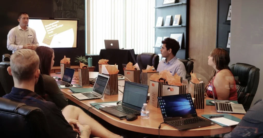 Seorang pembicara mempresentasikan materi kepada peserta rapat perusahaan yang duduk mengelilingi meja dengan laptop dan dokumen.